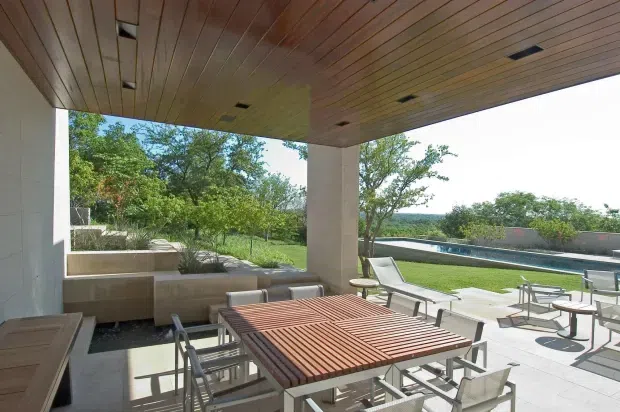 A covered outdoor dining and lounge area with a large slatted timber dining table, metal-framed chairs, built-in concrete seating, and a timber-lined ceiling. The open pavilion overlooks a landscaped garden and rectangular swimming pool, with clean modern lines and extensive outdoor views.