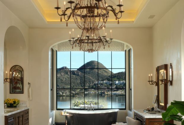 A richly decorated interior sitting area or landing with a dramatic floor-to-ceiling bay window overlooking a lake and mountainous landscape. The room features ornate wrought-iron chandeliers and wall sconces, cream plaster walls, arched openings, a recessed tray ceiling with warm concealed lighting, dark traditional cabinetry, and a curved upholstered seating or desk area positioned at the window.