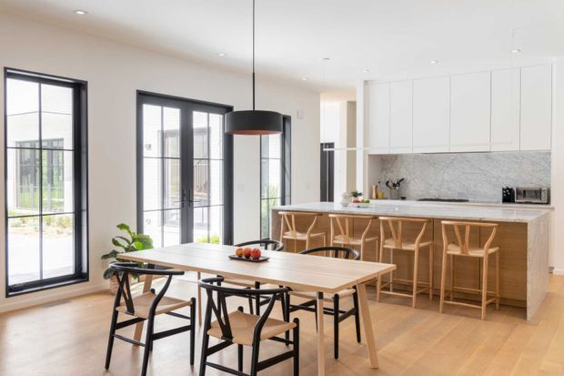 Bright, open-plan contemporary kitchen and dining area with pale timber flooring, white full-height cabinetry, a large island with timber bar stools, and a matching dining table with black-framed chairs. Tall black-framed glazed doors and windows bring in abundant natural light, while a dark pendant light provides contrast above the dining table.