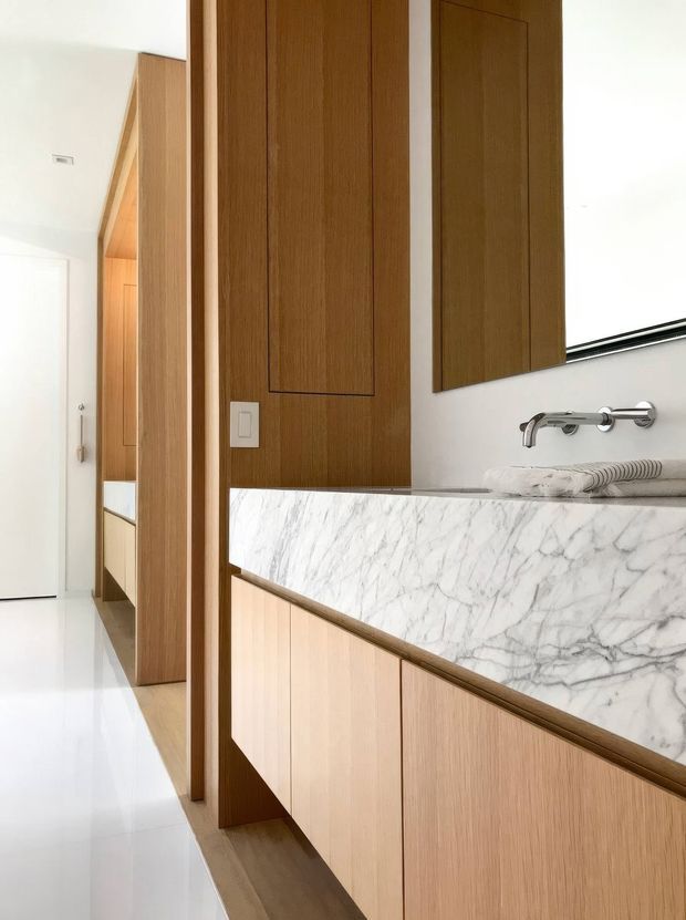 A refined contemporary bathroom vanity viewed from an adjacent hallway. The room features a long floating timber vanity with pale wood cabinetry, a thick white marble benchtop with prominent grey veining, wall-mounted chrome tapware, and matching vertical timber wall panelling and cabinet fronts. A bright white tiled or polished floor and a frameless mirror contribute to the clean, light-filled appearance.