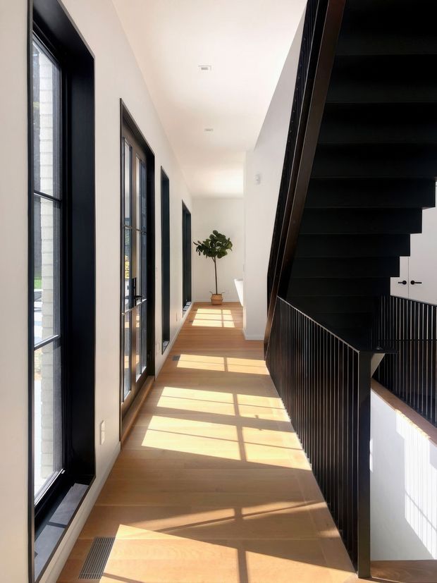 A bright, minimalist upper-level hallway with warm timber flooring, crisp white walls, black-framed windows and doors along one side, and a dramatic black stair with slender vertical balustrading on the other. Strong sunlight creates geometric shadows across the floor, while a potted tree adds a small natural accent at the end of the corridor.