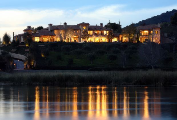 Dusk exterior view of a large, warmly illuminated stone-clad estate or resort-style residence on a landscaped hillside beside calm water. The building comprises multiple connected wings with varied pitched and hipped roof forms, chimneys, balconies, and many glowing windows. Reflections of the lights stretch across the water in the foreground, with wooded terrain behind.