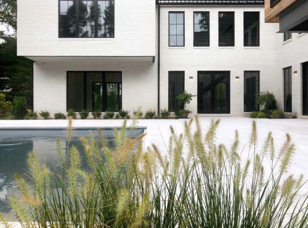 Contemporary multi-storey residence viewed from a landscaped pool area. The facade combines white brickwork, large black-framed windows and doors, and projecting upper-level volumes. A pale paved terrace surrounds a rectangular swimming pool, with ornamental grasses in the foreground and mature planting along the building.