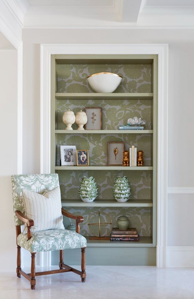 A refined interior vignette featuring a built-in recessed display bookcase with multiple shelves, framed by white decorative trim and filled with curated ornaments, books, framed photographs, ceramics, and greenery. The shelving has a muted sage-green floral patterned wallpaper backing. A traditional wooden armchair upholstered in coordinating botanical fabric sits in front, with white walls, ornate ceiling cornices, and pale flooring completing the elegant setting.