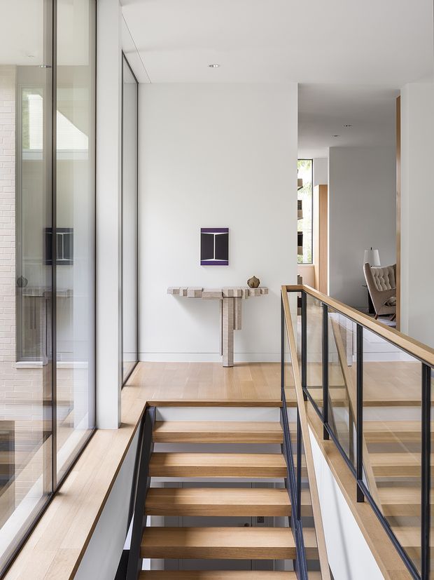 A bright contemporary interior landing viewed from a stairway, featuring open-riser timber stairs, slim black metal structure, and a glass balustrade with a timber handrail. Full-height glazing lines the left side, while a minimalist console table and small wall-mounted artwork occupy the white wall ahead. A sitting area is partially visible through the opening on the right.