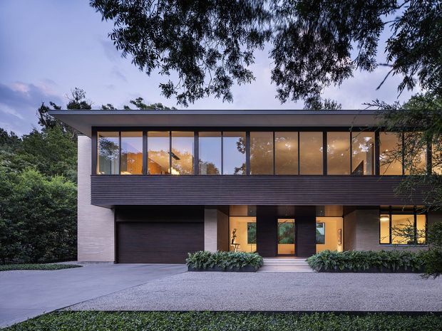 A two-storey contemporary residential facade photographed at dusk, set within mature landscaping. The house features a broad low-pitched roof, extensive upper-level glazing, dark horizontal cladding across the upper facade, pale masonry walls, a dark integrated garage, and a recessed entry with warm interior lighting.