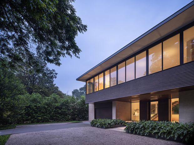 A dusk exterior view of a refined two-storey contemporary residence set among mature landscaping. The building features a long, low-pitched roof canopy, extensive upper-level floor-to-ceiling glazing, dark horizontal wall cladding, and a recessed ground floor with warm interior lighting. A paved driveway and dense planting frame the facade.