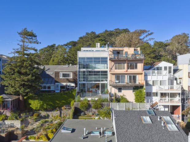 Exterior view of a dense residential neighbourhood on a wooded, sloping site. Several closely spaced multi-storey homes are visible, including a prominent contemporary house with a tall glazed facade and flat roof, alongside traditional and coastal-style residences. Rooftops, balconies, decks, landscaped gardens and retaining walls are visible under clear blue skies.