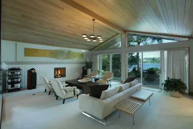 A spacious, light-filled mid-century-inspired living room with a low slatted timber ceiling, full-height glazing, and views toward a body of water. The room features a pale sectional sofa, sculptural lounge chairs, a fireplace set into a circular-patterned feature wall, a timber slatted coffee table, and a compact dining area near the windows. The overall atmosphere is refined, airy, and warm, with strong indoor-outdoor visual connection.