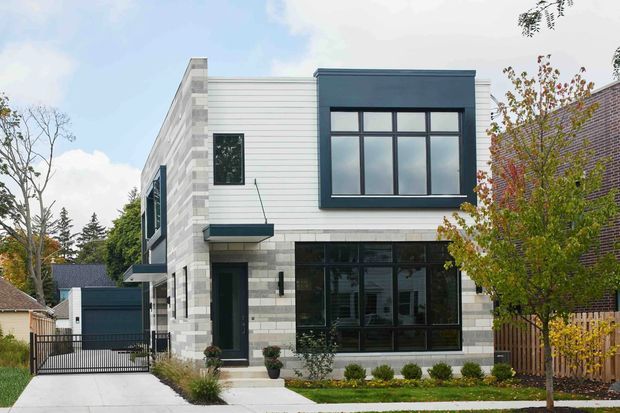 Exterior street-view photograph of a contemporary two-storey urban residence. The compact house has a flat roof, crisp geometric massing, pale horizontal cladding and light masonry blocks, with a dark charcoal projecting upper volume, large black-framed windows, a glazed ground-floor frontage, a small entry canopy, landscaped planting, and a gated driveway.