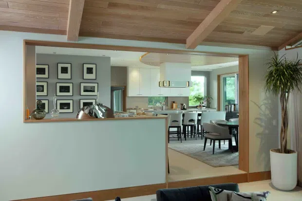Open-plan contemporary interior viewed from a living area toward a bright kitchen and dining space. A broad timber-framed opening and half-height partition define the transition, with white kitchen cabinetry, a central island, pendant lighting, and a dining table on a rug. The room features exposed timber ceiling boards and substantial timber beams, pale walls, timber flooring, framed artwork, and a tall indoor plant.