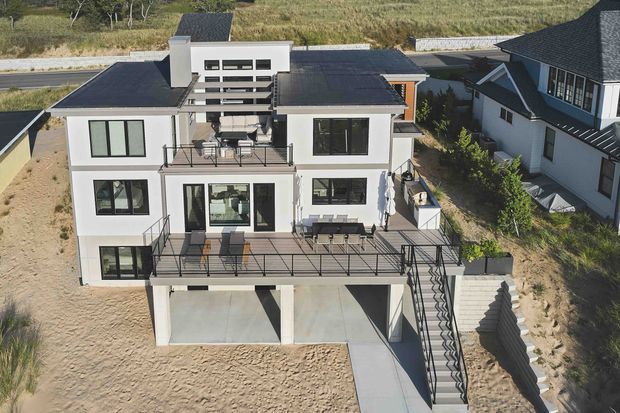 Aerial exterior view of a contemporary multi-storey house on a sloping sandy site. The white rendered facade has extensive black-framed glazing, several projecting balconies and terraces with dark metal balustrades, a flat and low-pitched roof composition, and an elevated lower level containing open parking or garage bays. A prominent external stair connects the upper terrace to the ground level. Adjacent modern homes and a rural landscape are visible in the background.