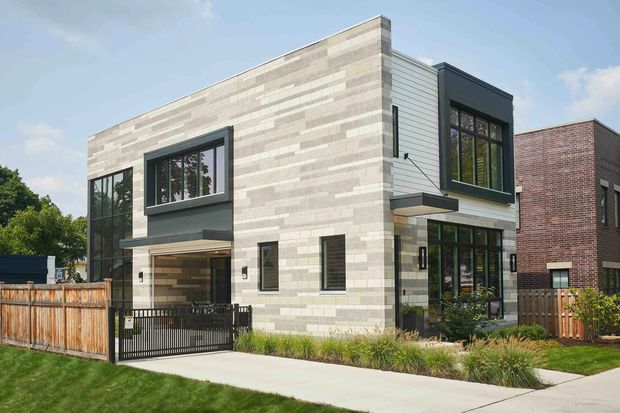 Exterior view of a contemporary two-storey urban residence with a rectilinear form, large dark-framed windows, projecting entry canopies, and a pale horizontally varied masonry facade. A timber privacy fence, landscaped planting, driveway gate, and concrete footpath frame the property, with a neighbouring brick building visible to the side.
