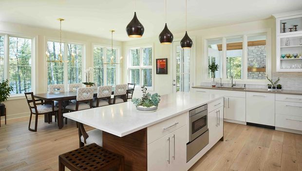Bright, spacious open-plan kitchen and dining area with a large white island, integrated stainless-steel appliances, white shaker-style cabinetry, light timber flooring, and dark pendant lights. A dining table sits beside expansive windows overlooking trees, while a servery window and sink are visible along the rear wall.