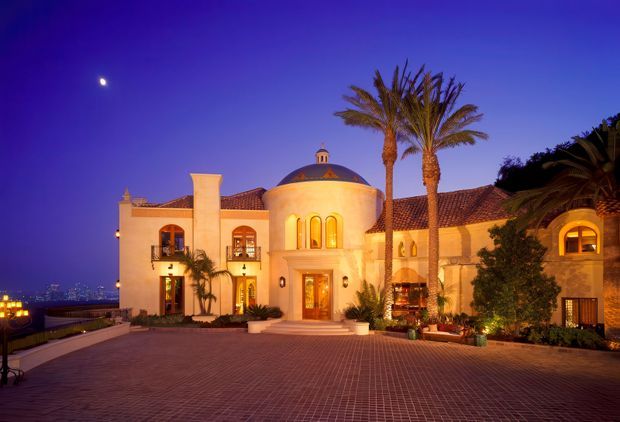 Night-time exterior view of a grand Mediterranean-style residence or estate, with a warm illuminated stucco facade, terracotta tiled roofs, a central domed entrance pavilion, arched windows and doors, balconies, tall palm trees, and a broad brick-paved forecourt. Distant city lights are visible beyond the property.