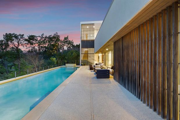 A contemporary multi-level residential exterior viewed from a long poolside terrace at dusk. The image shows a turquoise swimming pool, pale paved decking, a warm timber vertical-slat privacy screen, recessed exterior lighting, and a white geometric building with large glazed openings and a tall illuminated stair or circulation volume. Trees and a dusk sky form the background.