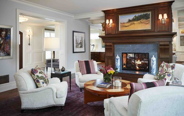 A refined traditional living room centered on a substantial dark timber mantel and fireplace with built-in cabinetry and an artwork panel above. The room contains upholstered armchairs and sofas, a round wooden coffee table, table lamps, floral cushions, framed art, and a deep purple rug. Pale grey walls, white trim, and an adjoining bright hallway reinforce the classic, elegant character.
