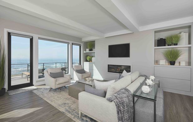 Bright contemporary living room with floor-to-ceiling glazed doors and windows opening to a balcony with an ocean view. The room features a large light sectional sofa, armchairs, glass coffee table, wall-mounted television, linear fireplace, built-in shelving and cabinetry, layered area rug, and dark wood-look flooring. A recessed tray-style ceiling adds architectural definition.