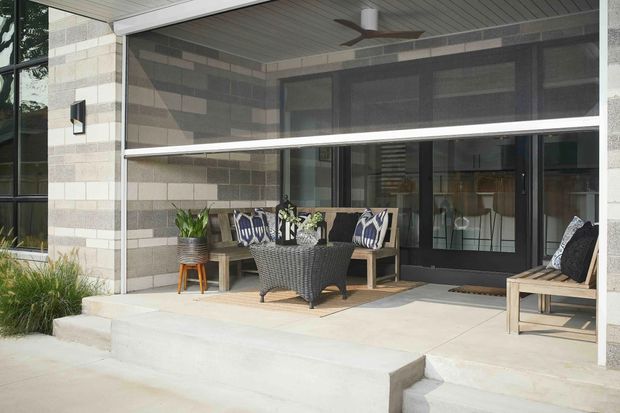 A covered outdoor terrace or porch adjoining a contemporary home, with a concrete slab and steps, dark-framed glazed doors and windows, light masonry walls, a slatted ceiling with ceiling fan, and wicker lounge furniture arranged around a small table. Potted plants and cushions add a relaxed residential outdoor setting.