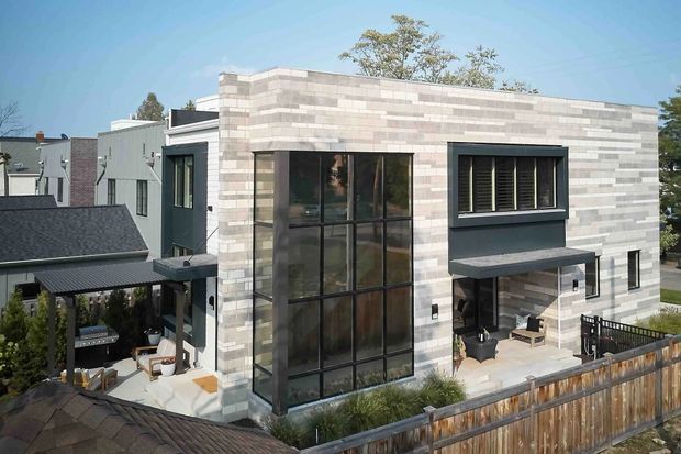 Exterior view of a contemporary multi-level residence with a flat roof and a pale horizontally coursed masonry or stone-clad facade. The building features extensive black-framed floor-to-ceiling glazing, a projecting dark canopy over the entry or outdoor terrace, and a fenced landscaped courtyard or patio. Adjacent residential buildings are visible in the background.