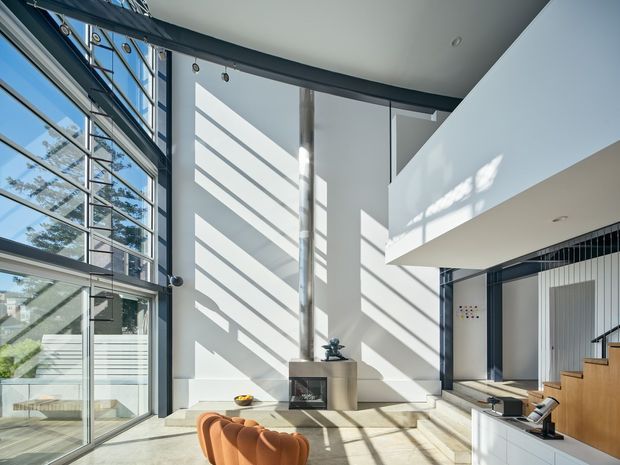 Bright double-height contemporary living space with expansive floor-to-ceiling glazing, a sculptural fireplace wall, mezzanine-like upper volume, and a timber staircase. Strong linear sunlight casts dramatic shadows across the pale walls and concrete-toned floor. A partial sculptural lounge chair and built-in cabinetry are visible in the foreground.