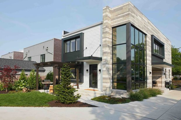 Contemporary multi-storey residential facade with a prominent two-storey corner of floor-to-ceiling glazing, pale horizontal cladding, and light stacked-stone masonry. The home features dark-framed windows, a recessed entry, a covered side terrace or garage-like opening, landscaped garden beds, and a broad concrete driveway or walkway. Neighbouring residential buildings are visible in the background.
