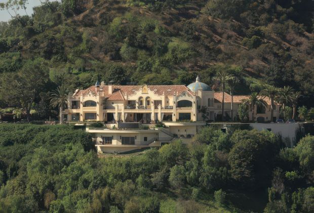 Exterior view of a large ornate multi-storey villa set into a densely vegetated, sloping hillside. The building has cream stucco walls, extensive arched openings and balconies, terracotta tiled roofs, a prominent small domed turret, and landscaped grounds with mature palms and shrubs.