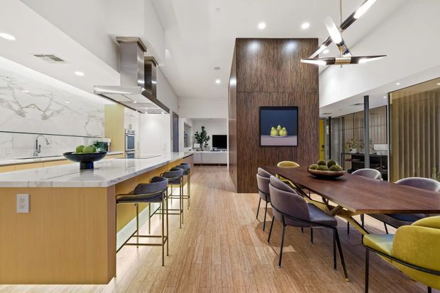 Open-plan contemporary kitchen and dining area with a large light-wood island, pale stone benchtop, integrated stainless-steel appliances and a marble-look splashback. A dark timber feature wall separates the kitchen from the dining space, which has a long dark wood table, upholstered dining chairs, and modern pendant and track lighting. Light timber flooring and white walls create a bright, refined interior.