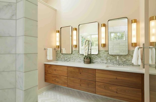 Bright contemporary bathroom featuring a long floating timber vanity with multiple drawers, a pale stone benchtop and twin integrated basins. The vanity wall has a mosaic tile splashback, three rounded rectangular mirrors, and warm brass-and-glass wall sconces. Pale geometric floor and wall tiles create a clean, refined finish, with a tiled shower enclosure visible at left.