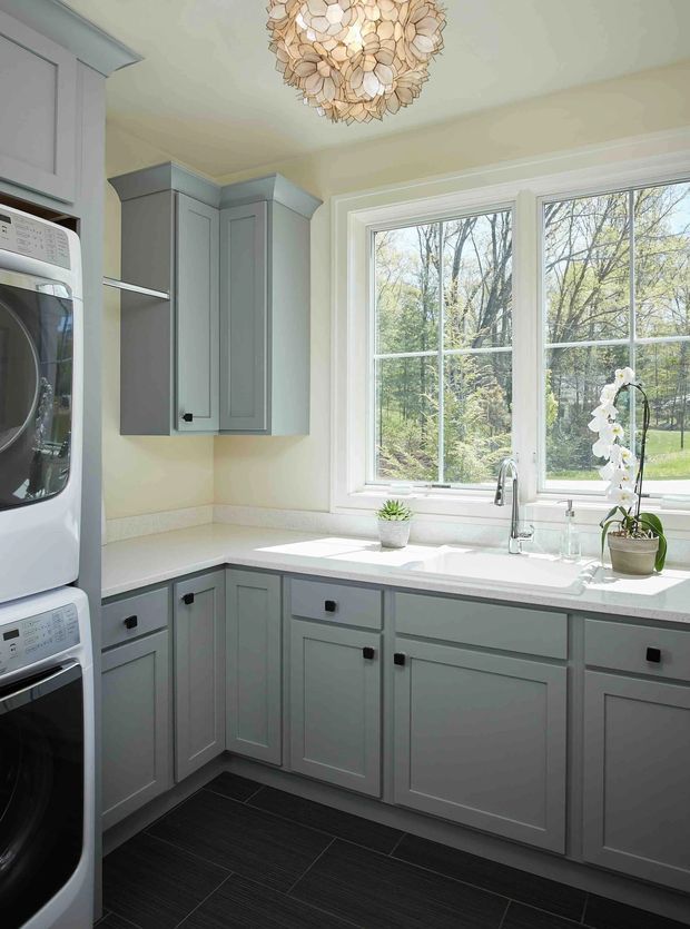 Bright, compact laundry room with pale grey shaker-style cabinetry arranged in an L-shaped layout, a white countertop and integrated sink beneath a large double window. Stacked white washer and dryer occupy the left side, while a decorative floral pendant light and potted orchids add character. The dark tiled floor contrasts with the light cabinetry and walls, and the room overlooks a leafy outdoor setting.