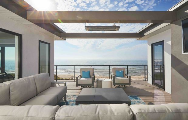 A bright contemporary coastal outdoor living area opening directly onto a timber deck overlooking the ocean. The space features a large light-coloured sectional sofa, lounge chairs with blue cushions, a coffee table, dark structural beams, extensive glazing, and a partially open pergola-style roof with a skylight. A balustrade frames the beachfront view.