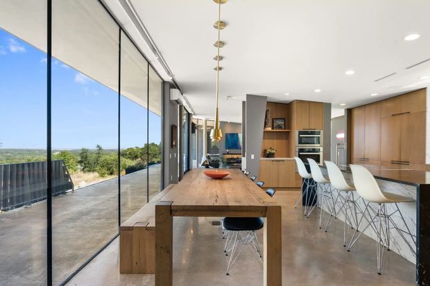 Bright open-plan dining and kitchen interior with a long solid timber dining table, contemporary white bar stools, warm timber cabinetry, integrated appliances, polished concrete flooring, and full-height black-framed glazing opening to a broad outdoor view. A linear cluster of brass pendant lights hangs above the table, while a living area is visible beyond the kitchen.