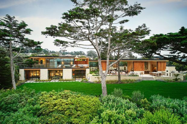 Wide exterior view of a contemporary, low-rise residence set within extensive landscaped gardens. The house has a horizontally composed form with large glazed openings, warm timber-clad sections, pale masonry walls, flat or very low-pitched rooflines, terraces, and stepped outdoor areas. Mature trees and dense planting frame the building, with a broad lawn in the foreground.