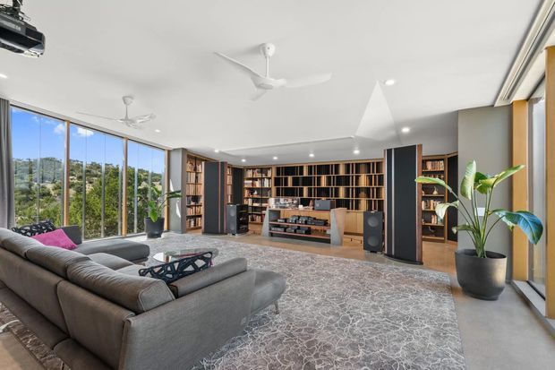 A spacious, light-filled contemporary living room with floor-to-ceiling windows overlooking greenery. The room features a large grey sectional sofa, patterned area rug, low sculptural coffee table, recessed lighting, ceiling fans, and an extensive dark timber built-in library and media wall with open shelving and integrated cabinetry. Large potted plants and warm timber accents soften the refined entertainment space.