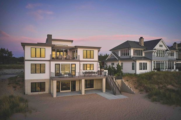 Exterior architectural view of two neighbouring multi-storey homes at dusk. The main home is a contemporary, flat-roofed structure with pale rendered walls, extensive dark-framed glazing, multiple balconies and terraces, and an external stair descending to a sandy landscaped foreground. A larger traditional gabled house with light weatherboard cladding is visible alongside it.