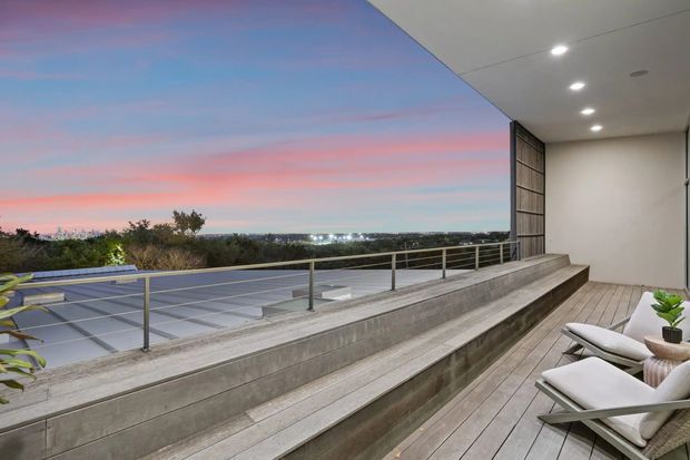 A contemporary elevated outdoor terrace at dusk, overlooking a distant suburban or coastal landscape. The terrace features broad stepped concrete seating or retaining walls, timber-look decking, slim metal balustrades, recessed ceiling downlights, and a large open roofed section. Two pale lounge chairs and potted greenery sit along the right side, while a flat roof or adjoining structure extends across the left foreground.
