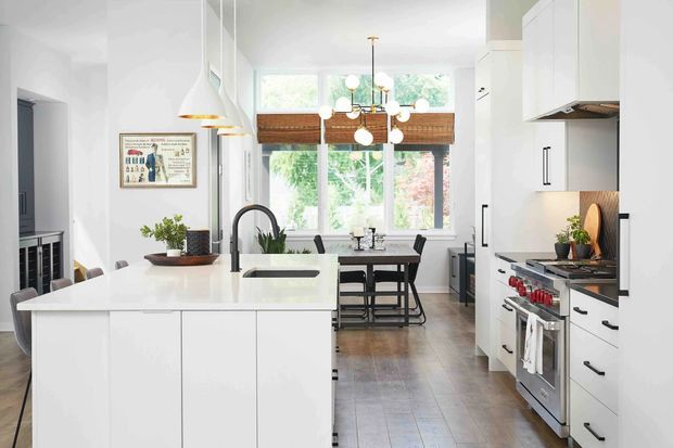 Bright contemporary kitchen with a large white island, integrated sink and black tap, white flat-panel cabinetry, stainless-steel range, tiled floor, and a dining area positioned in front of large windows with timber blinds. A linear pendant and globe chandelier provide feature lighting, while a partial living area is visible at left.