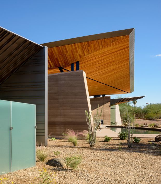 Exterior architectural photograph of a contemporary, angular building in a dry desert landscape. The composition features dramatic cantilevered roof planes, warm timber soffits, dark metal wall cladding, textured earth-toned walls, and frosted glass doors. Gravel landscaping with sparse drought-tolerant planting surrounds the building, with a long narrow reflecting pool visible at right.