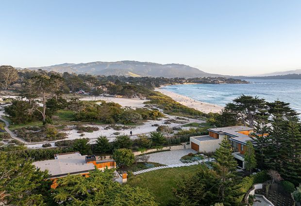 Aerial view of a coastal residential property overlooking a sandy beach and bay, with low modern homes set among established trees and landscaped gardens. Mountainous terrain is visible across the water, creating a scenic beachfront setting.