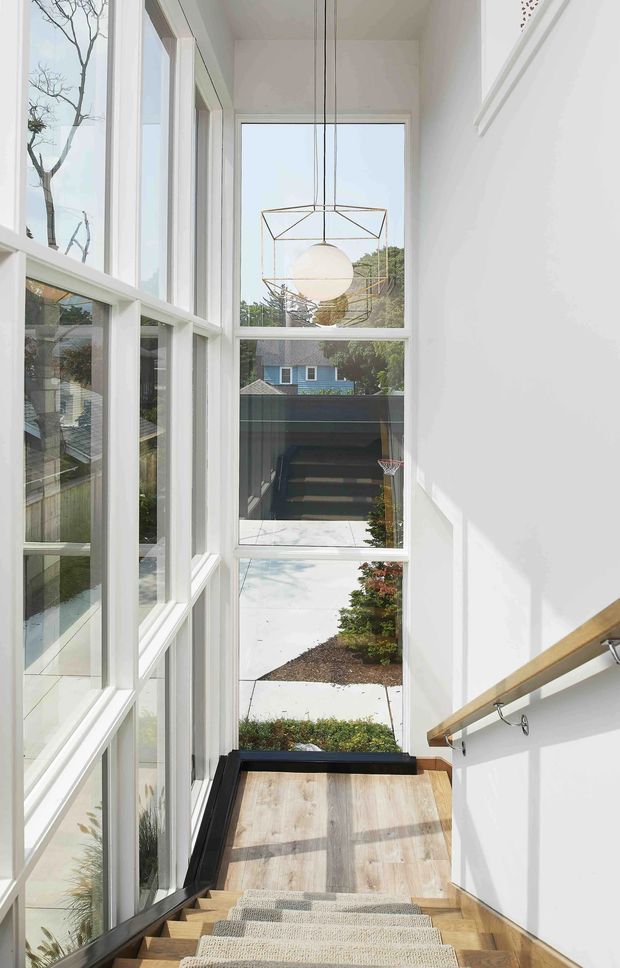 A bright, narrow stairway with a timber staircase and runner, white walls, a simple timber handrail, and extensive floor-to-ceiling glazing along the landing. A geometric pendant light hangs in front of the tall windows, which overlook a residential garden and neighbouring homes. The space is airy and contemporary with strong natural light and clean detailing.