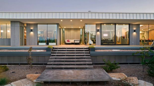 Front exterior view of a contemporary single-storey home with extensive floor-to-ceiling glazing, dark vertical facade panels, a broad low-pitched roof, and a raised terrace accessed by wide timber steps. Warm interior lighting and visible living spaces show through the glass, while minimalist landscaping and a concrete approach frame the entrance.