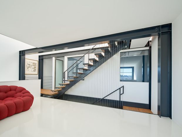 Bright contemporary interior view focused on an open stairway framed by dark metal architectural elements. The stair has timber treads, slender vertical metal balusters, and a dark handrail, connecting a lower and upper level. White walls, ceiling, and glossy white surfaces create a clean backdrop, with a partial sculptural red sofa in the foreground and glimpses of adjoining rooms and exterior views through glazing.