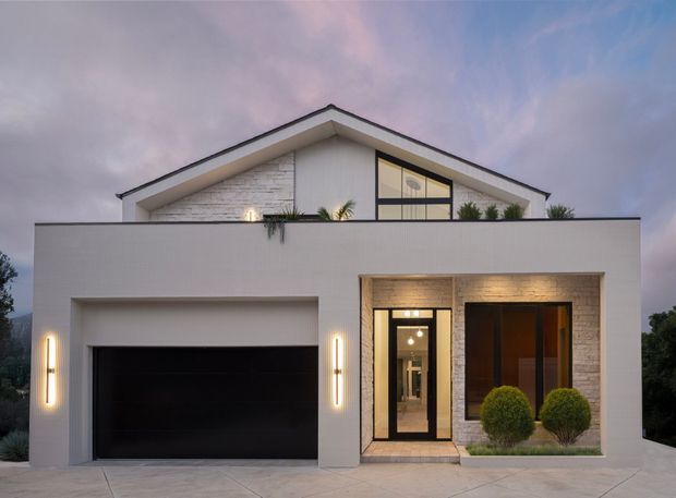 Contemporary two-storey suburban house facade at dusk, featuring a prominent pitched gable roof, white rendered walls, pale brick feature cladding, a large black garage door, warm vertical exterior lighting, and a recessed glazed entry framed by dark timber or metal. Upper-level glazing and a rooftop terrace or balcony planter area are visible behind the front parapet.