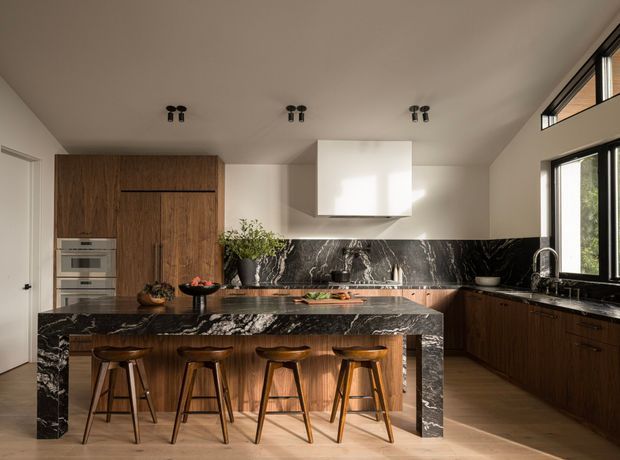 A contemporary kitchen with a large waterfall-edge island clad in dramatic dark veined stone, seating four timber bar stools. The room features dark timber cabinetry, integrated stainless-steel appliances, a matching stone splashback and benchtops, black-framed windows, and a sculptural white rangehood. Warm natural light enters from the windows and clerestory glazing.