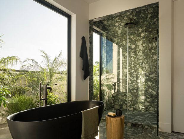 A sophisticated bathroom featuring a freestanding matte-black bathtub beside expansive glazing, with lush palms visible outside. The walk-in shower is enclosed by clear glass and fully clad in dramatic green natural stone with prominent pale veining and textured variation. A small timber stool and dark towel add warmth and contrast. The space is bright, refined, and strongly connected to the landscape.
