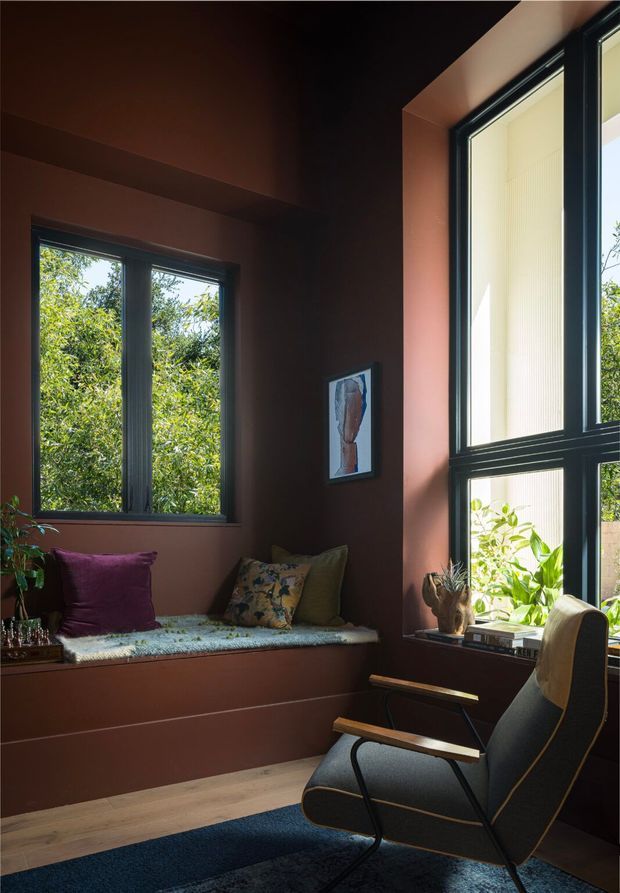 A warm, compact reading nook or sitting room with deep terracotta-brown walls, large black-framed windows, a built-in window seat with cushions, and a dark upholstered lounge chair. Natural greenery is visible outside and in a small indoor planter, while a framed artwork and books add understated decoration. The room has strong natural light, timber flooring, and a dark blue rug.