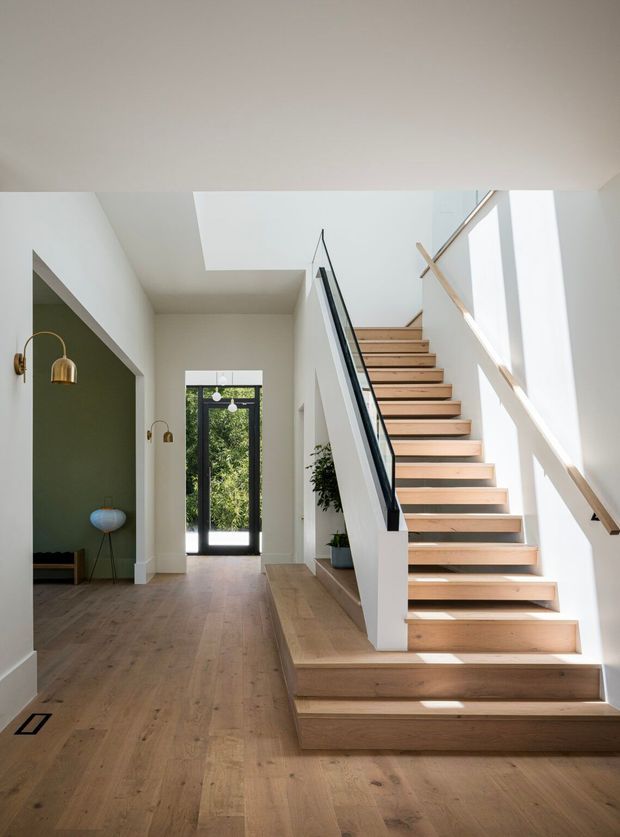 Bright contemporary residential entry hall dominated by a broad open timber staircase. The stair has pale wood treads and matching timber flooring, crisp white walls, a black slender balustrade, and a timber handrail. A large roof skylight brings daylight into the circulation space, while a glazed black-framed entry door and a recessed sitting area with pendant lights are visible at the rear and left.
