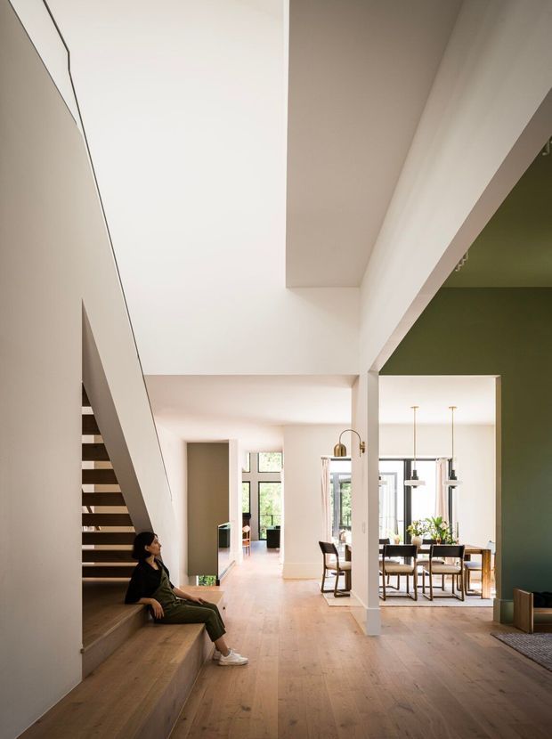 A bright, open-plan residential interior with a sculptural open staircase and integrated timber seating at left. The space has pale timber flooring, white walls and ceilings, a double-height void, and a dining area visible beyond with pendant lights and large glazed openings. A deep olive-green accent wall frames the right side, while a person sits on the stair bench. The overall design is clean, contemporary, and spacious with strong architectural lines.
