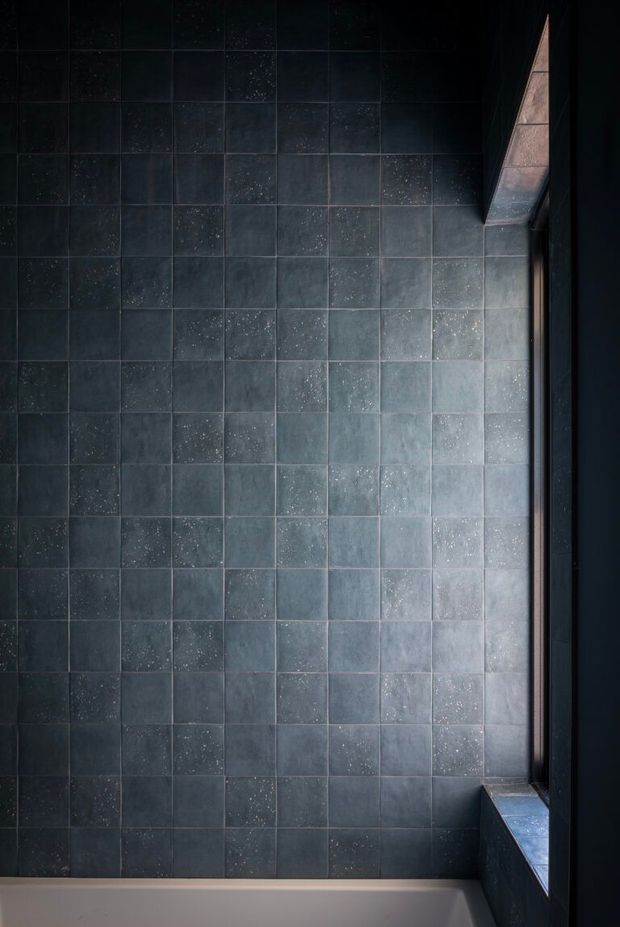 Close-up view of a dark, moody bathroom wall clad in square blue-grey ceramic or porcelain tiles, with subtle speckled variation and dark grout. A partial white bathtub is visible along the bottom, beside a deep-set window or recessed opening with dark framing and a tiled sill.