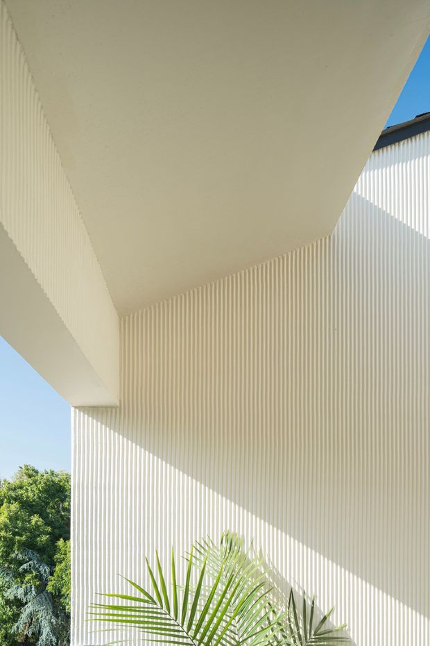 Close-up exterior architectural view of a pale ribbed facade beneath a broad projecting flat canopy. Strong sunlight creates crisp diagonal shadows across the vertically corrugated wall, with blue sky and landscaping visible at the edges. The image emphasises geometric lines, texture, and a clean contemporary building envelope.